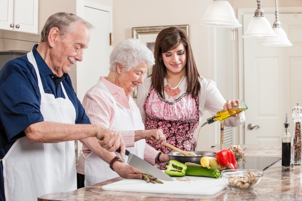 A young woman (granddaughter or caregiver) helps a senior couple prepare a meal in their residential kitchen.