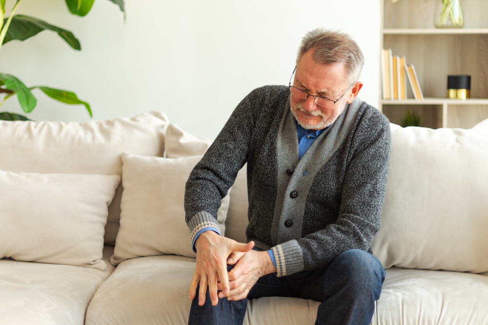 Knee pain arthritis body sick health care concept. Unhappy middle aged senior man suffering from knee ache sitting on sofa at home, Mature old senior grandfather touching leg feeling pain hurt in knee